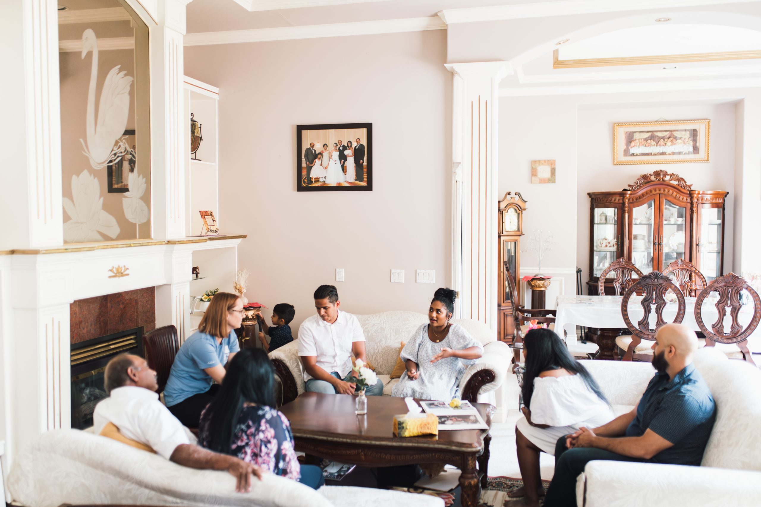 A diverse group of adults and children are gathered in an elegant living room with classical decor. The room is bathed in natural light, highlighting its off-white walls, a white fireplace with a decorative swan motif, and traditional furniture including a carved wooden cabinet and dining set. Several framed photographs adorn the walls, adding a personal touch to the space. The group is engaged in casual conversation facilitated by a Canuck Place counsellor. They are seated on sofas and a chair around a wooden coffee table 