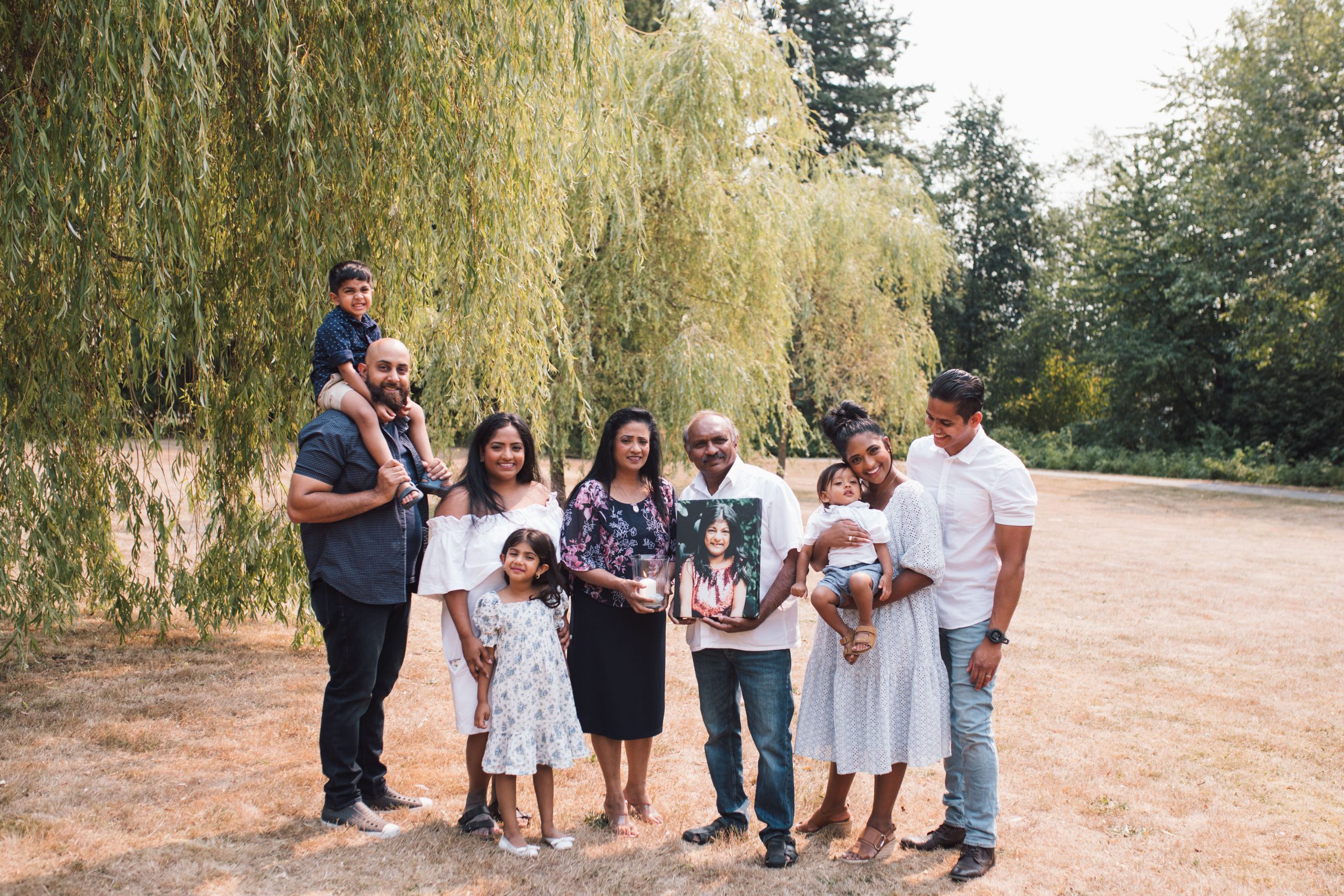 A family portrait is taken outdoors with a backdrop of a willow tree and a lush, leafy environment. To the left, a man joyfully carries a young boy on his shoulders. Centered in the photo is a smiling woman holding a candle in a glass jar, flanked by an older gentleman to her right who is holding a framed picture of a young girl. A woman to the right holds a baby, standing close to a man in a white shirt. In front of them, two young girls smile brightly. The family is dressed in a mixture of casual and dressy attire, creating a sense of a special occasion being celebrated informally. The overall mood is one of happiness and togetherness.