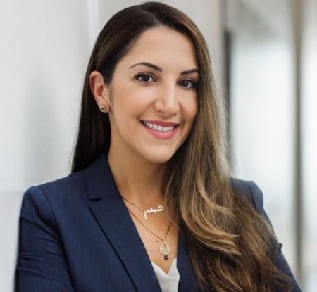 Portrait of a professional woman smiling at the camera. She has long, dark hair and is wearing a navy blue suit with a light blouse. She is wearing a silver necklace and black stud earrings. The background is a softly blurred office setting.