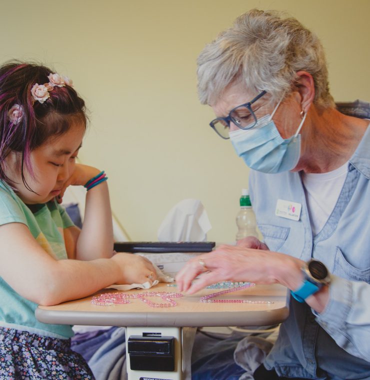 A young girl with downcast eyes is seated at a hospital bedside tray, with a Canuck Place family volunteer wearing a light blue shirt and a protective mask engaged in an activity with her. The worker is guiding the girl’s hands in a bead sorting or craft project and they both seem focused on the task at hand.