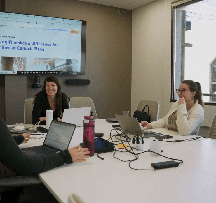 A group of four Canuck Place staff members engaged in a collaborative meeting around a conference table, filled with laptops and work materials. One woman is turned towards the camera, smiling warmly. A presentation is visible on a screen in the background. The room is well-lit with natural light streaming in from windows.