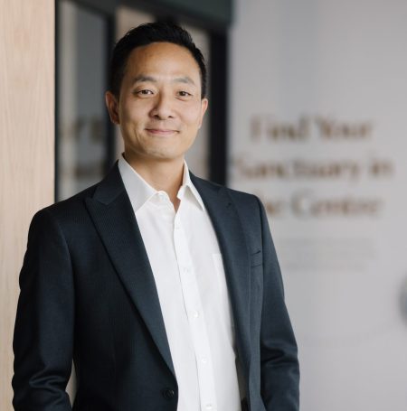 A professional portrait of a man standing in an office setting, dressed in a dark suit, white shirt, and brown belt. He smiles gently at the camera, positioned in front of a subtle background that includes the phrase 'Find Your Sanctuary in Canuck Centre'.