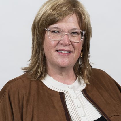 Professional headshot of a woman with blonde hair wearing clear glasses, a white button-up shirt, and a brown jacket, smiling against a plain white background.